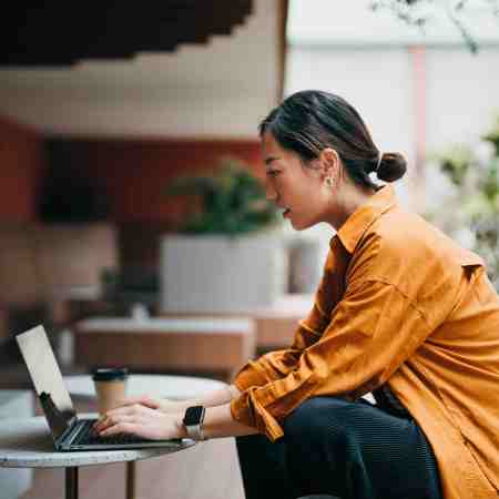 A woman in an orange shirt works on a laptop at an outdoor table.