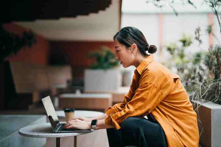A woman in an orange shirt works on a laptop at an outdoor table.